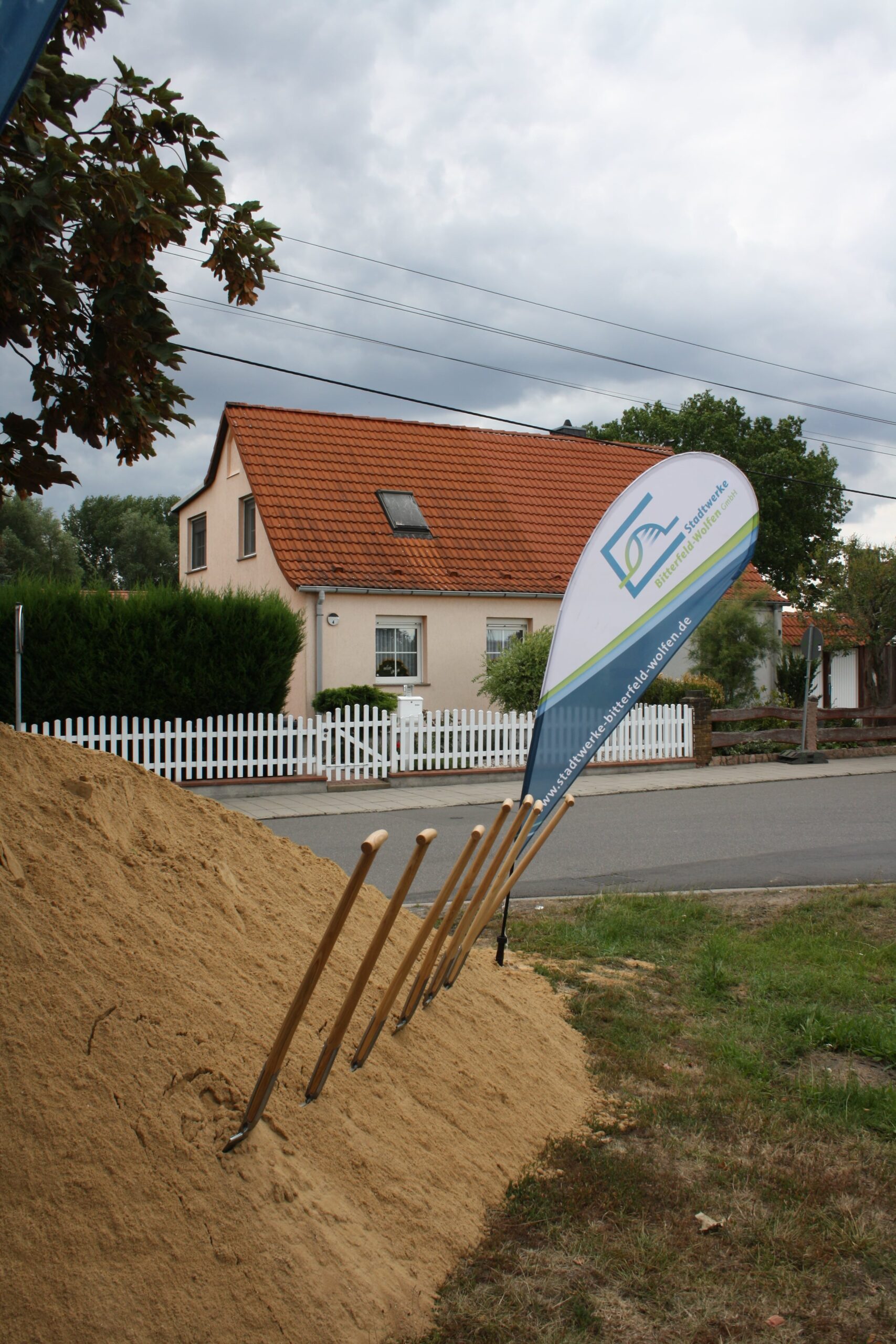 Ein Sandhaufen, in den mehrere Schaufeln gesteckt sind, steht an einer Straße; ein tropfenförmiges Schild und ein weißer Zaun mit einem Haus und Bäumen sind im Hintergrund unter einem bewölkten Himmel zu sehen.