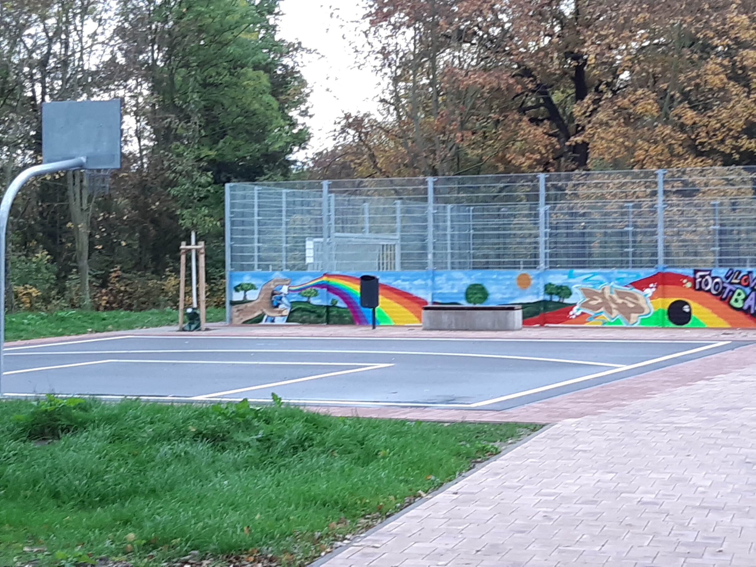 Ein kleiner Basketballplatz im Freien mit einem Wandgemälde an der Wand dahinter, das einen Regenbogen, Tiere und bunte Texte zeigt. Der Bereich ist von Bäumen und Grünflächen umgeben, mit einem gepflasterten Gehweg im Vordergrund.