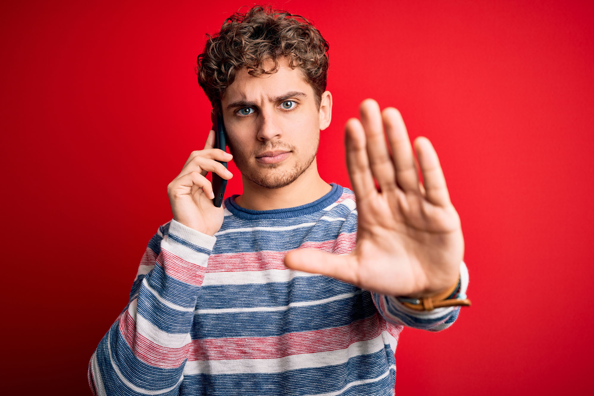 Ein junger Mann mit lockigem Haar und einem gestreiften Pullover hält sich ein Telefon ans Ohr und hebt die andere Hand in einer Stopp-Geste, während er vor einem leuchtend roten Hintergrund steht.